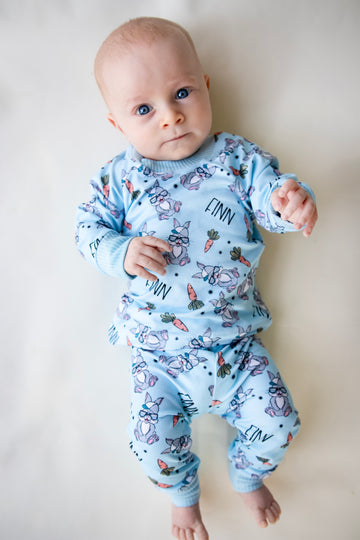 baby boy wearing boys easter personalized pajamas featuring his name, hip easter bunnies in glasses and hats, and cartoon carrots on light blue stretchy knit fabric. 