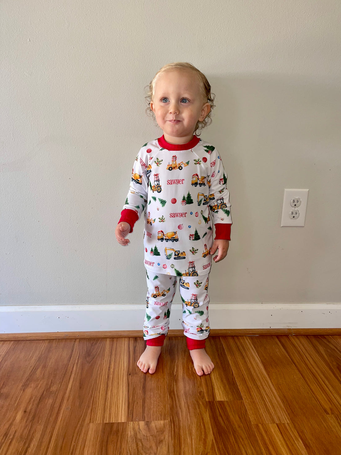 little boy wearing Christmas Construction custom holiday pajamas with construction vehicles in santa hats and christmas trees, featuring child's name in red letters and red cuffs.