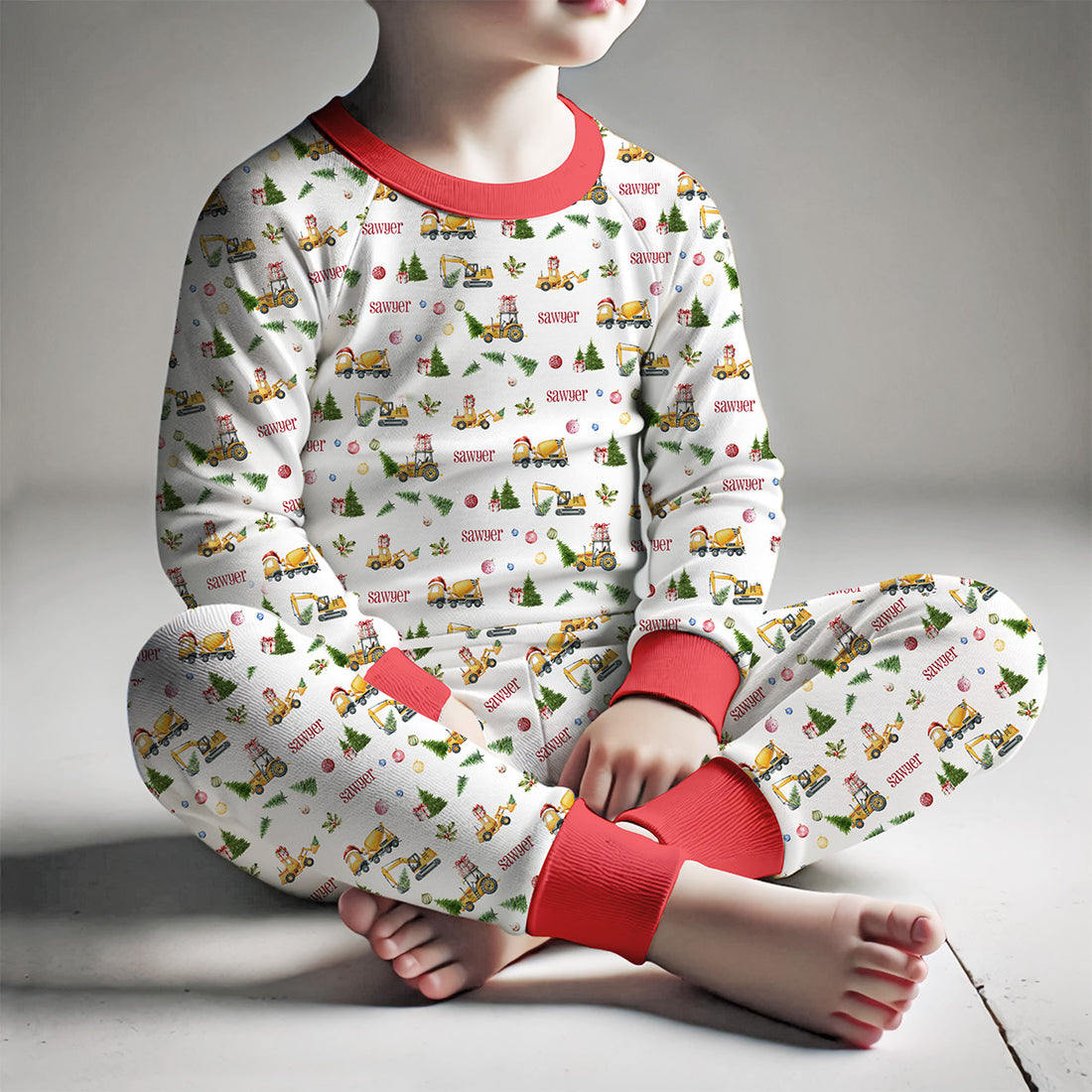 little boy wearing Christmas Construction custom holiday pajamas with construction vehicles in santa hats and christmas trees, featuring child's name in red letters and red cuffs.