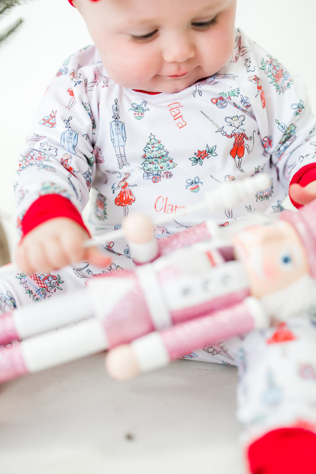 baby wearing personalized Modern Nutcracker Ballet pajamas, featuring minimalistic images from the iconic nutcracker ballet among child's name on white fabric with red cuffs. 