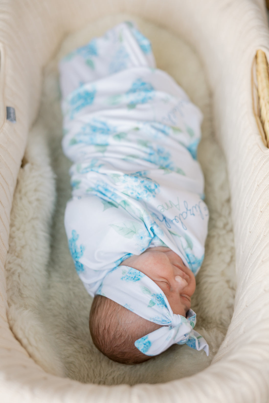 Newborn baby swaddled in a floral blanket with watercolor blue hydrangea print and baby's name on a white background. Matching headband