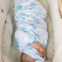 Newborn baby swaddled in a floral blanket with watercolor blue hydrangea print and baby's name on a white background. Matching headband