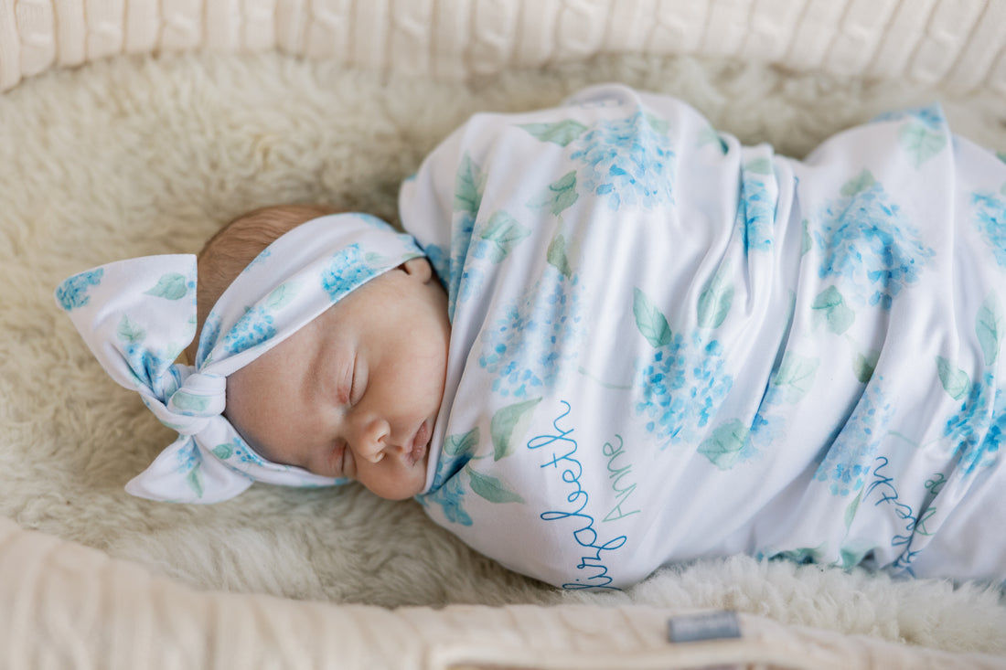 Newborn baby swaddled in a floral blanket with watercolor blue hydrangea print and baby's name on a white background. Matching headband