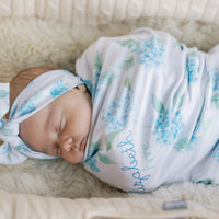 Newborn baby swaddled in a floral blanket with watercolor blue hydrangea print and baby's name on a white background. Matching headband