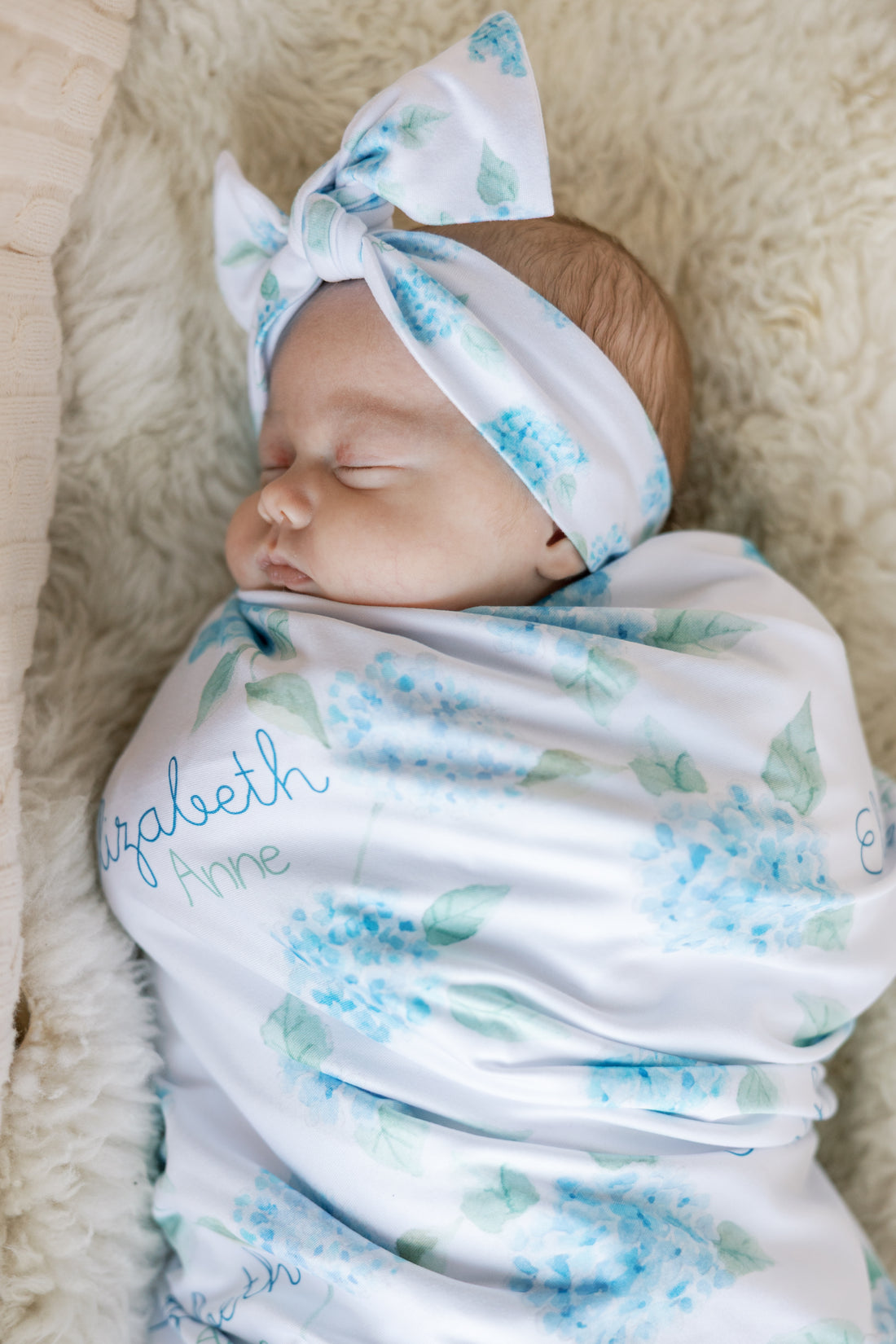 Newborn baby swaddled in a floral blanket with watercolor blue hydrangea print and baby's name on a white background. Matching headband