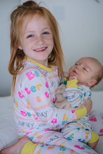 Big sister cuddling baby sibling in matching personalized pajamas with colorful letters and names, showing soft stretchy fabric and cozy comfort.