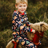 Little boy wearing personalized Dino Cookie print Christmas pajamas, navy blue background with dinosaur shaped cookies wearing Christmas sweaters