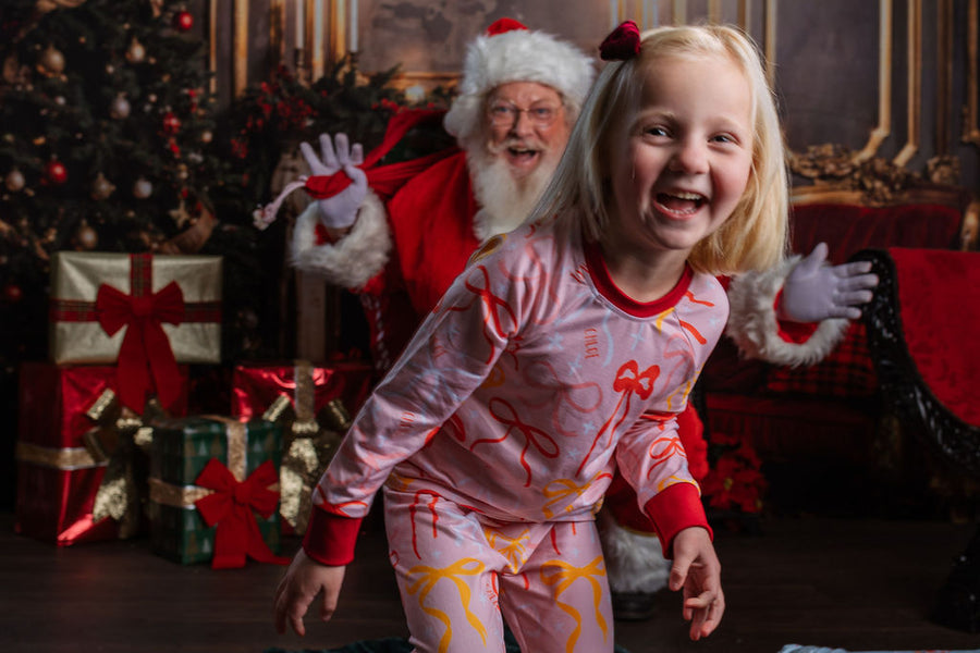 Little girl wearing pink personalized christmas pajamas with red pink and orange bows, and her name in red letters scattered around with red cuffs