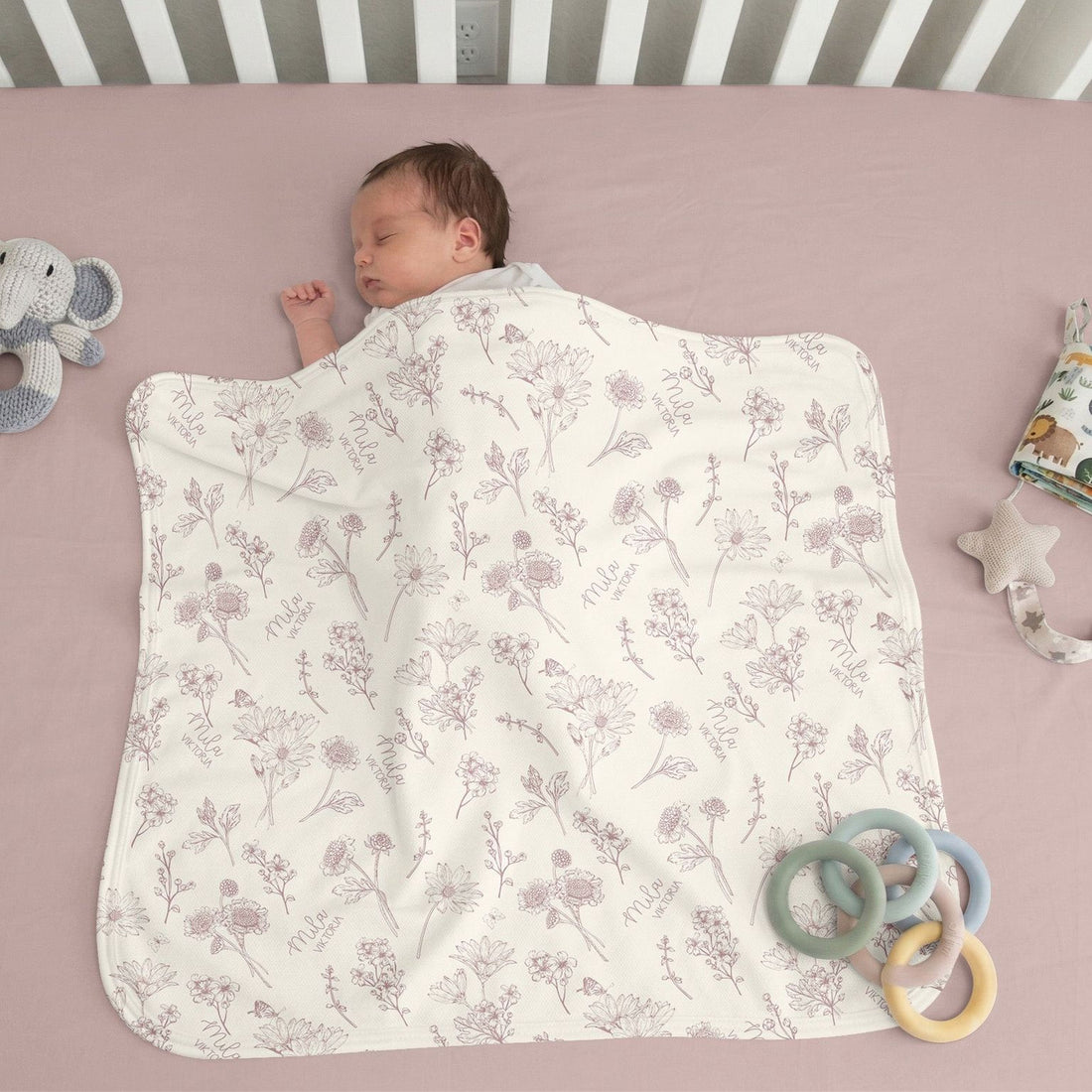 A sleeping baby swaddled in a blanket with a floral pattern is lying on a pink bedspread.