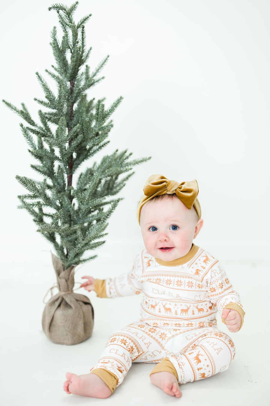 Little baby sitting in front of a Christmas tree wearing yellow striped Christmas pajamas. 