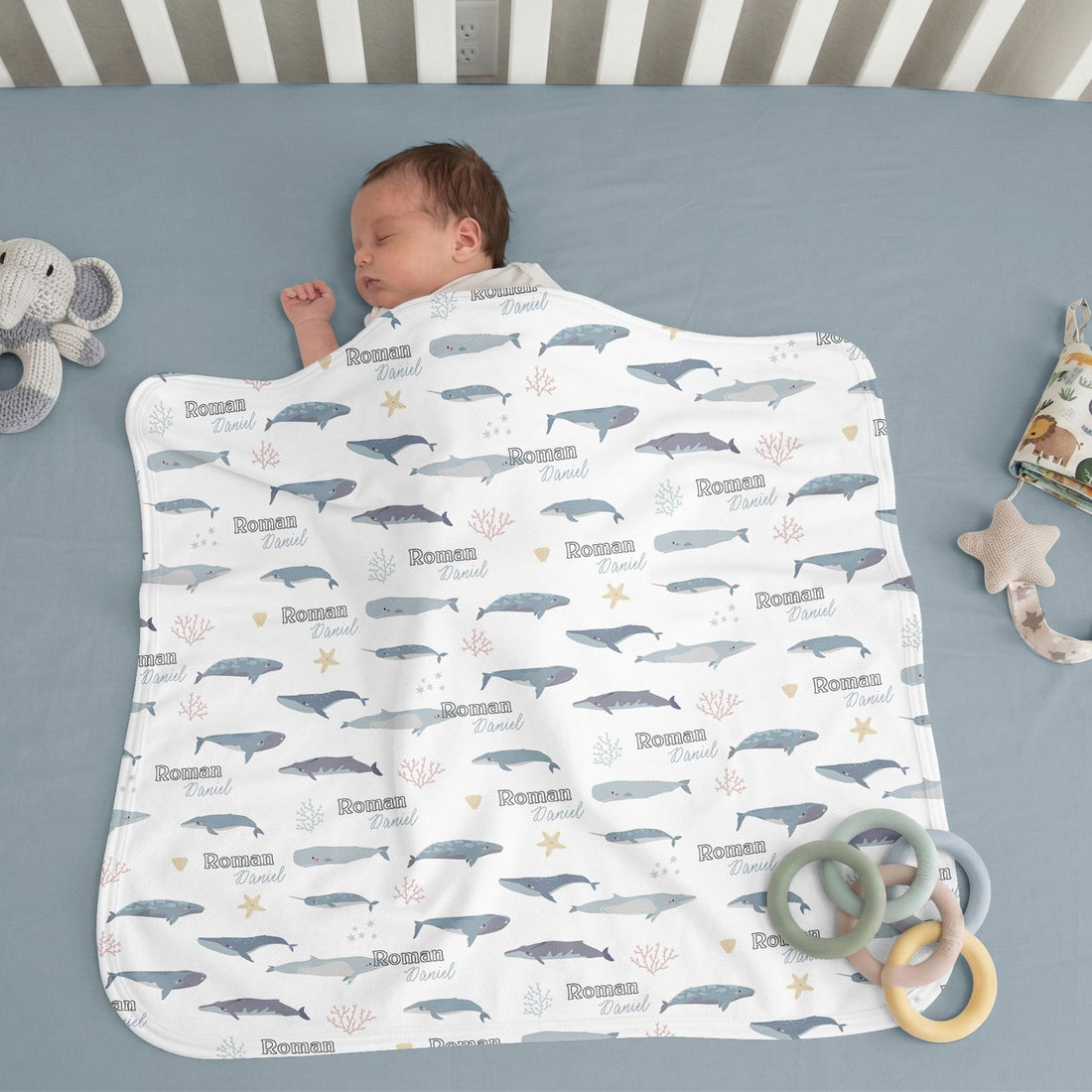 A baby is sleeping peacefully on a blue blanket with a whale and starfish pattern, surrounded by toys.