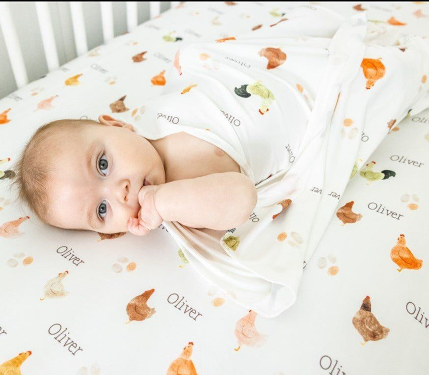 A baby is lying on a white bed with a blanket featuring a chicken pattern. The baby is looking at the camera with a curious expression.