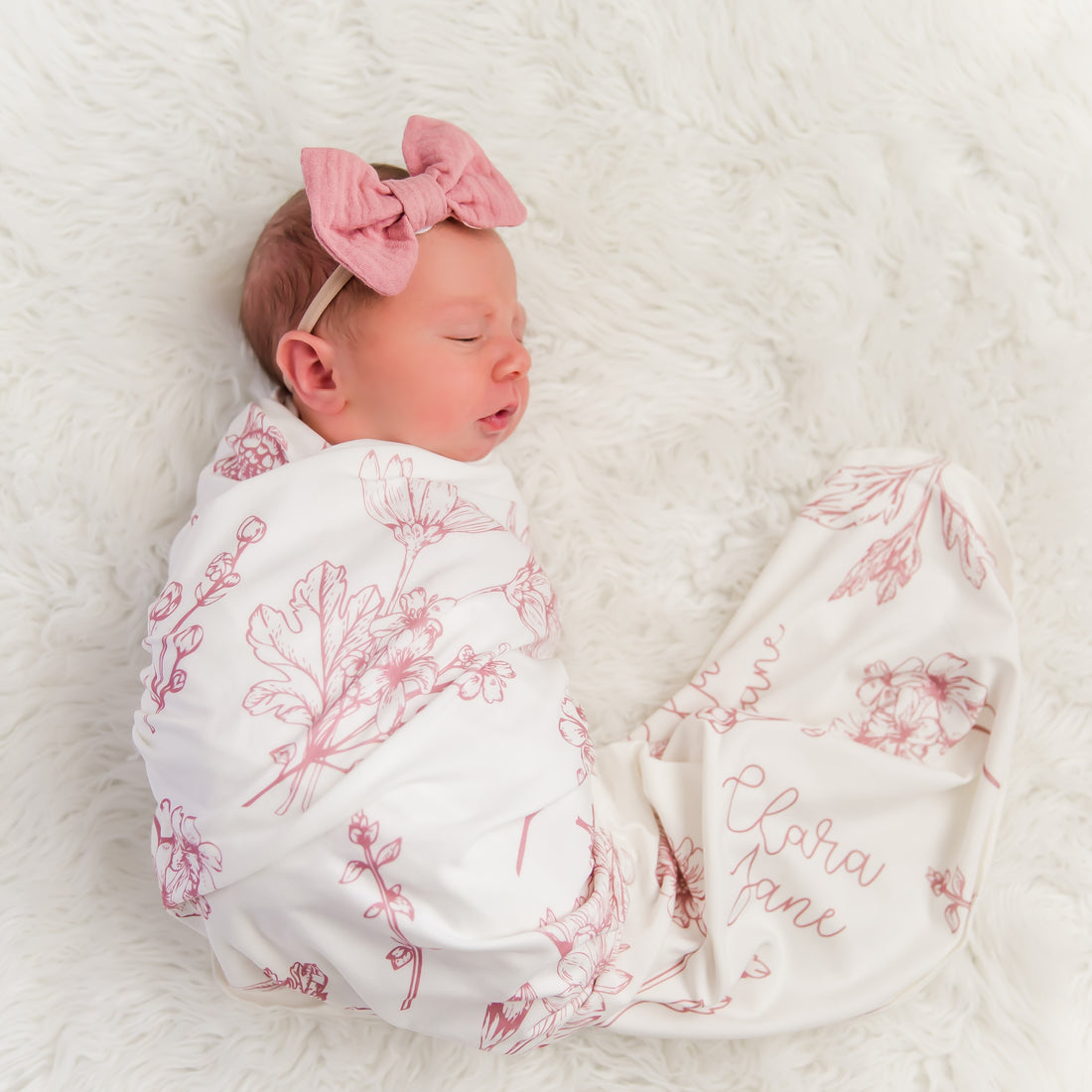 A newborn baby girl is peacefully sleeping, swaddled in a white blanket with a pink floral pattern and wearing a pink bow.