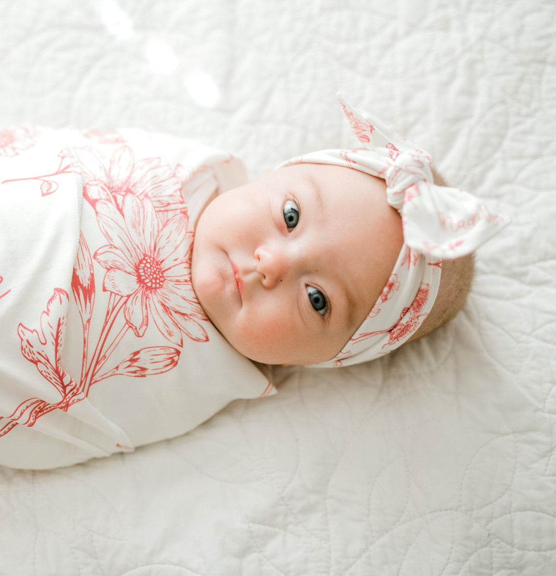 A baby girl is lying on a white bed, wearing a white floral blanket and a white headband with red flowers.