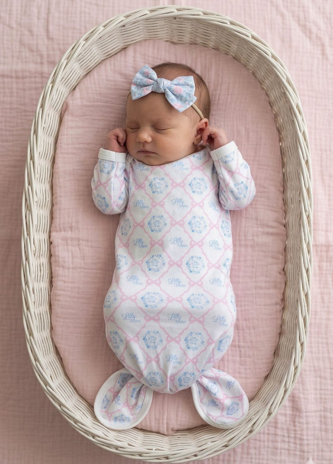 A newborn baby girl is peacefully sleeping in a white wicker basket, wearing a pink and white patterned onesie and a blue bow.