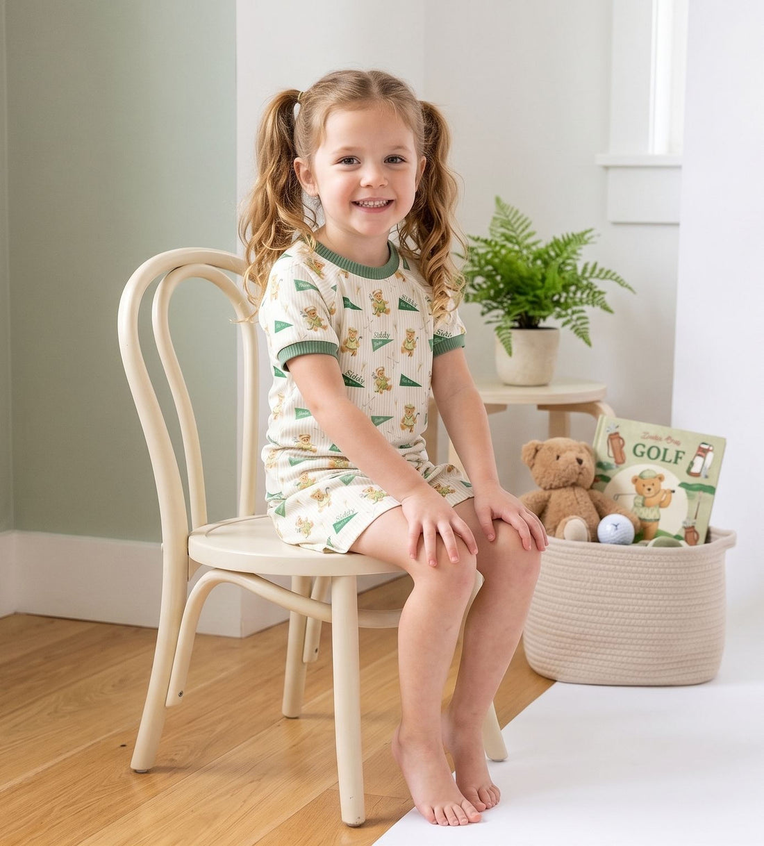 A young girl in a green and white pajama top and shorts is sitting on a white chair, smiling at the camera. Next to her on the floor is a basket filled with toys, including a teddy bear and a golf ball.