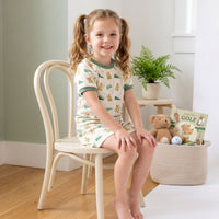 A young girl in a green and white pajama top and shorts is sitting on a white chair, smiling at the camera. Next to her on the floor is a basket filled with toys, including a teddy bear and a golf ball.