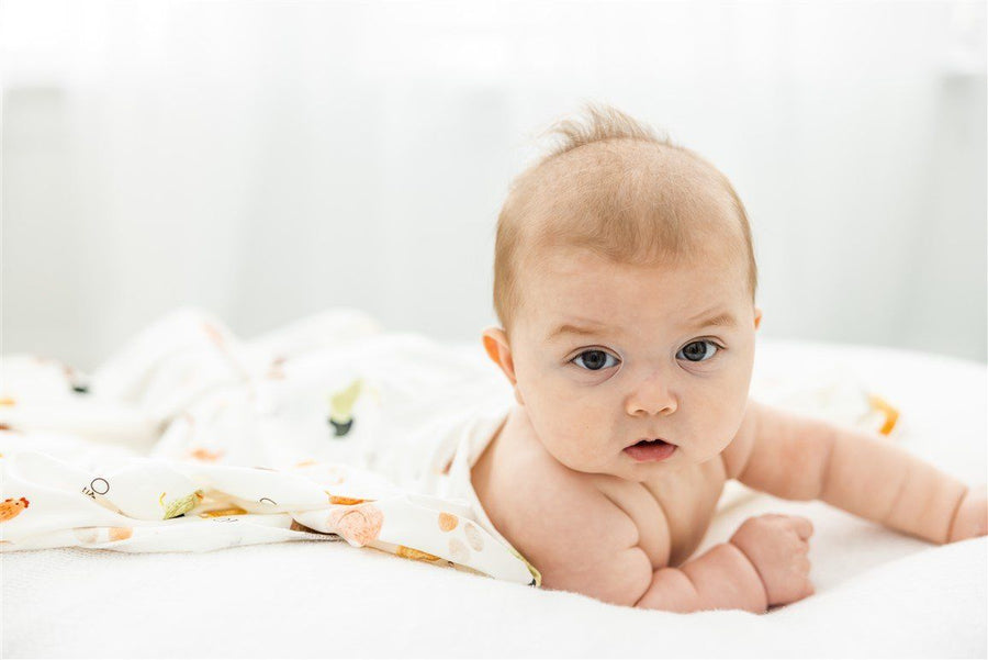 A baby lying on a white surface, looking directly at the camera with a curious expression.