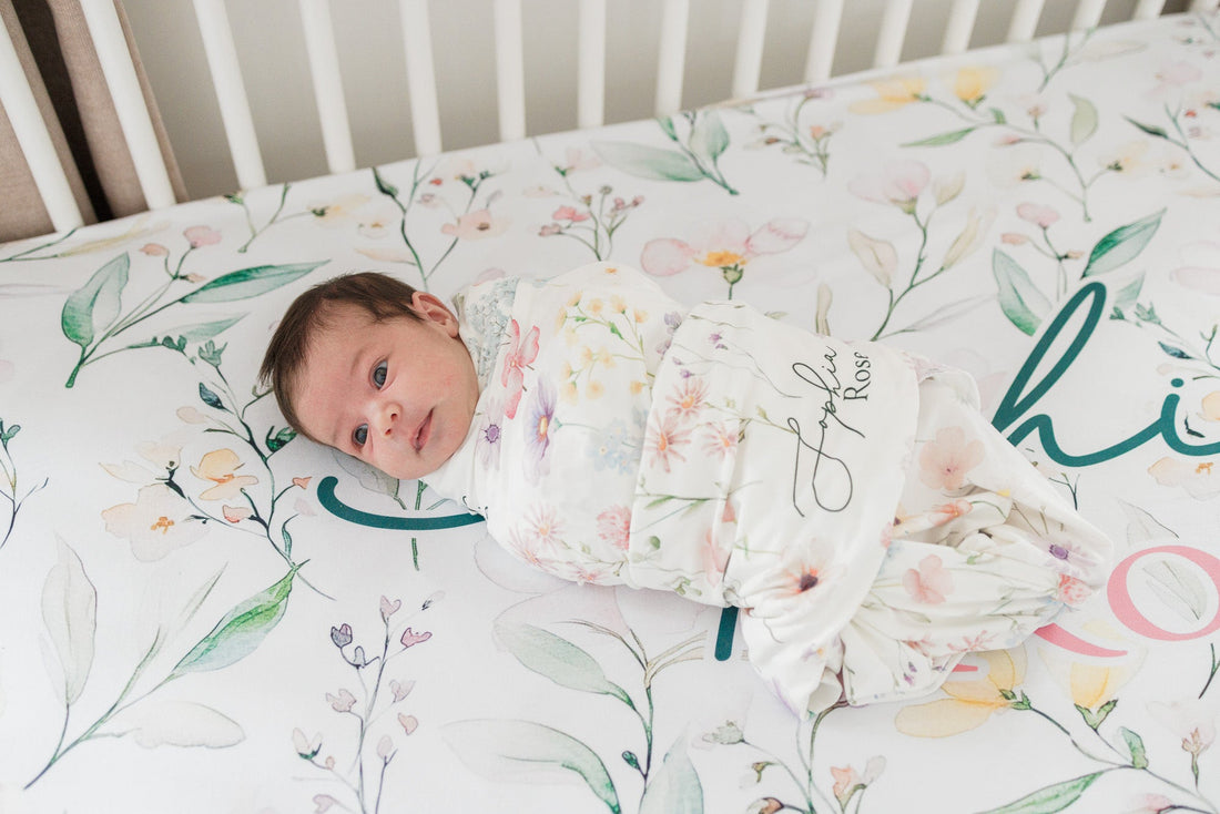 A newborn baby is swaddled in a floral blanket and lying on a white bed with a floral pattern.