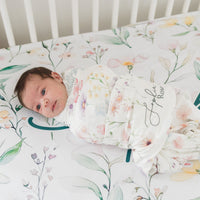 A newborn baby is swaddled in a floral blanket and lying on a white bed with a floral pattern.