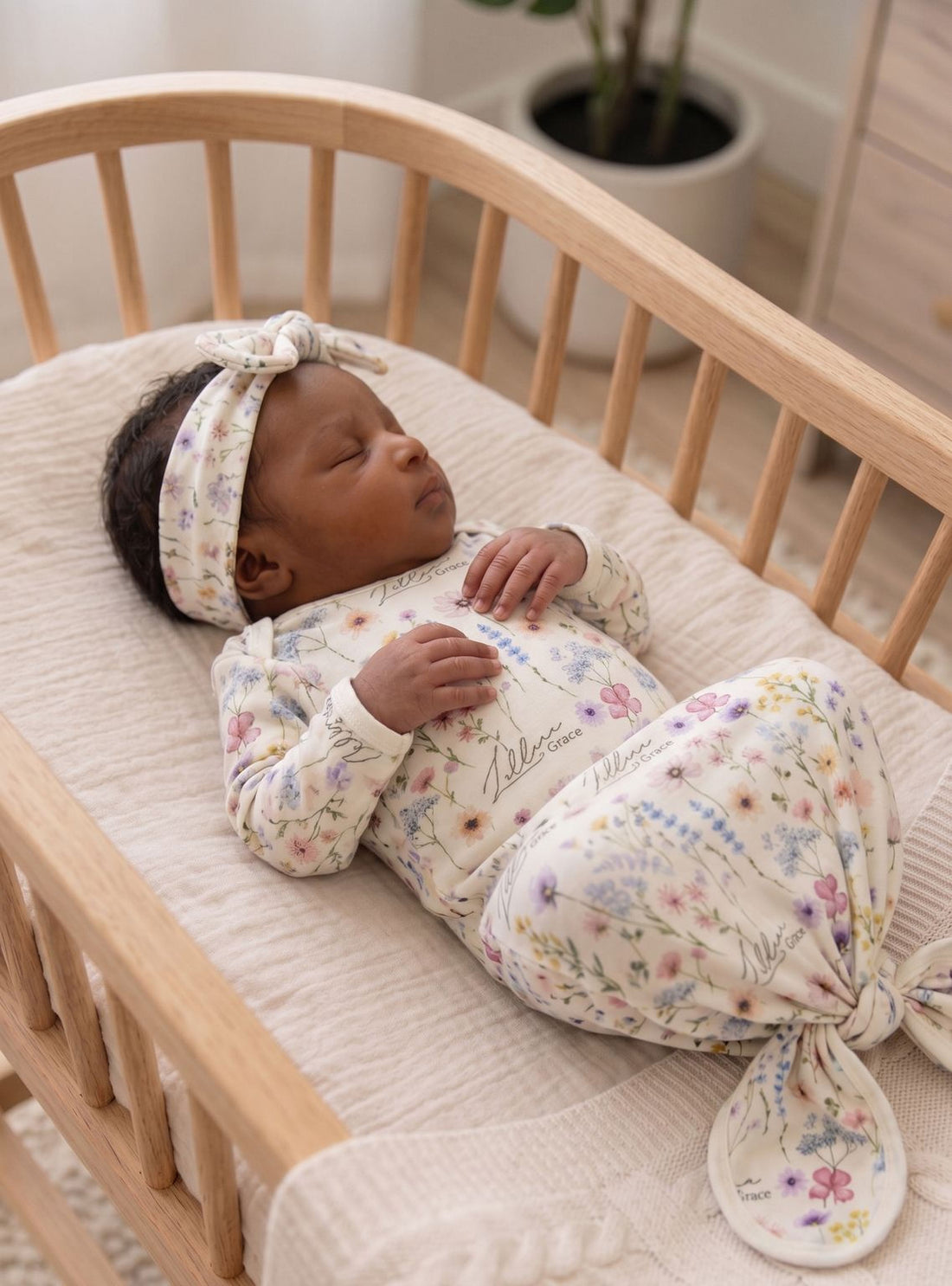 A newborn baby girl is peacefully sleeping in a wooden crib, wearing a white floral onesie and a matching headband.