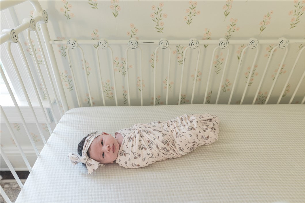 A baby swaddled in a floral print blanket is lying on a white mattress, looking at the camera.