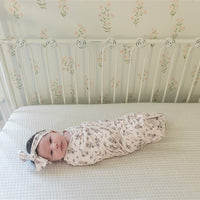 A baby swaddled in a floral print blanket is lying on a white mattress, looking at the camera.