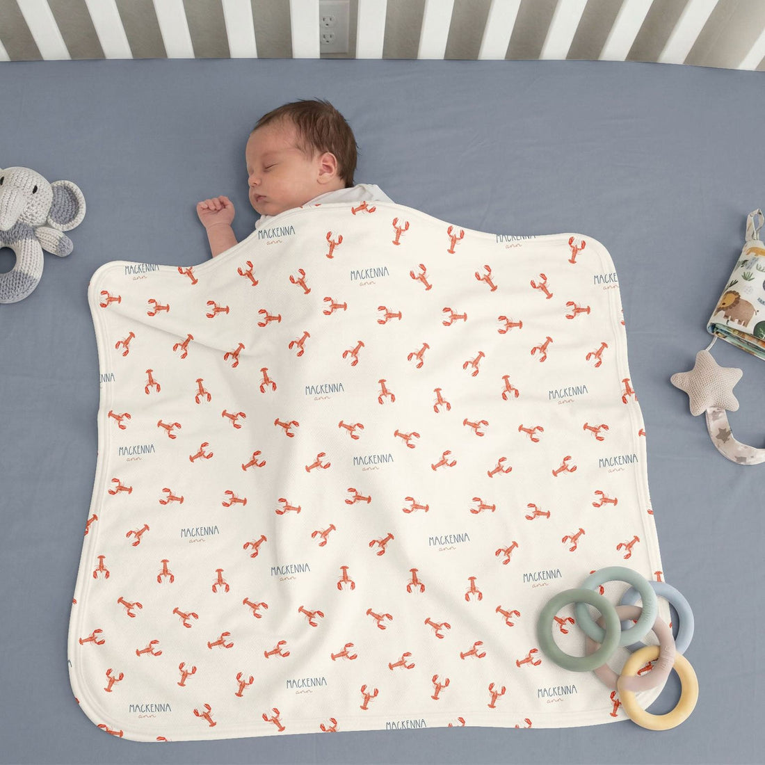 A sleeping baby wrapped in a blanket with a lobster pattern, surrounded by toys on a blue surface.
