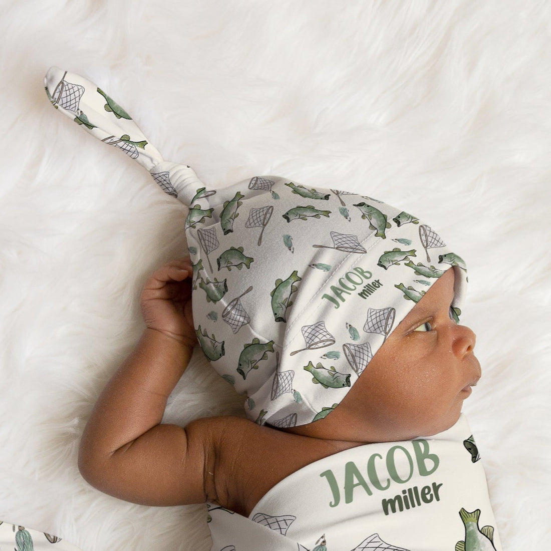 Newborn baby wrapped in a fish-themed swaddle and hat on a white fur background