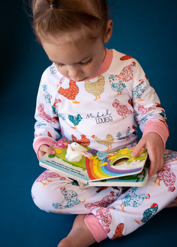 little girl wearing personalized chicken pajamas with a calico design in various colors, her name in script font on a white knit background, and pink cuffs. 