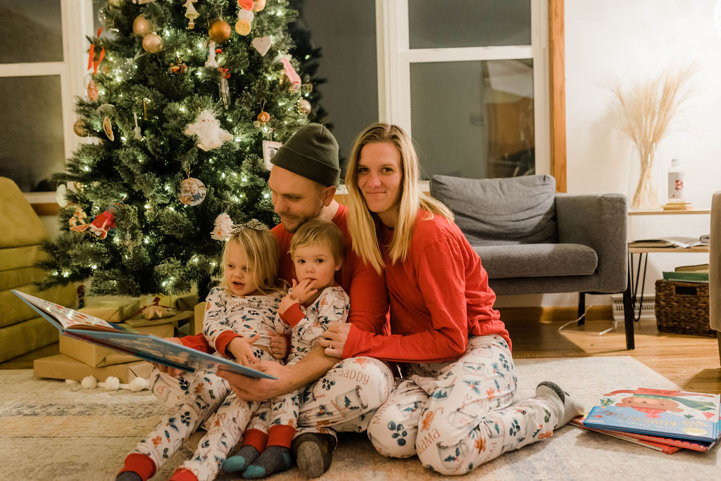 Family wearing matching Hot Cocoa Christmas Pajamas and reading a book in front of the Christmas tree. 