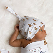 Baby wrapped in a white blanket with teddy bear pattern and baby's name in  text, lying on a soft white surface.
