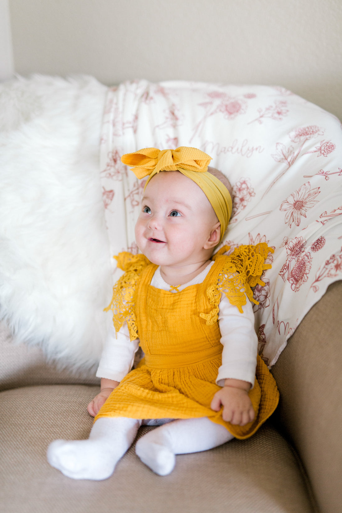A baby girl wearing a yellow dress and a yellow headband is sitting on a couch, looking up and smiling.