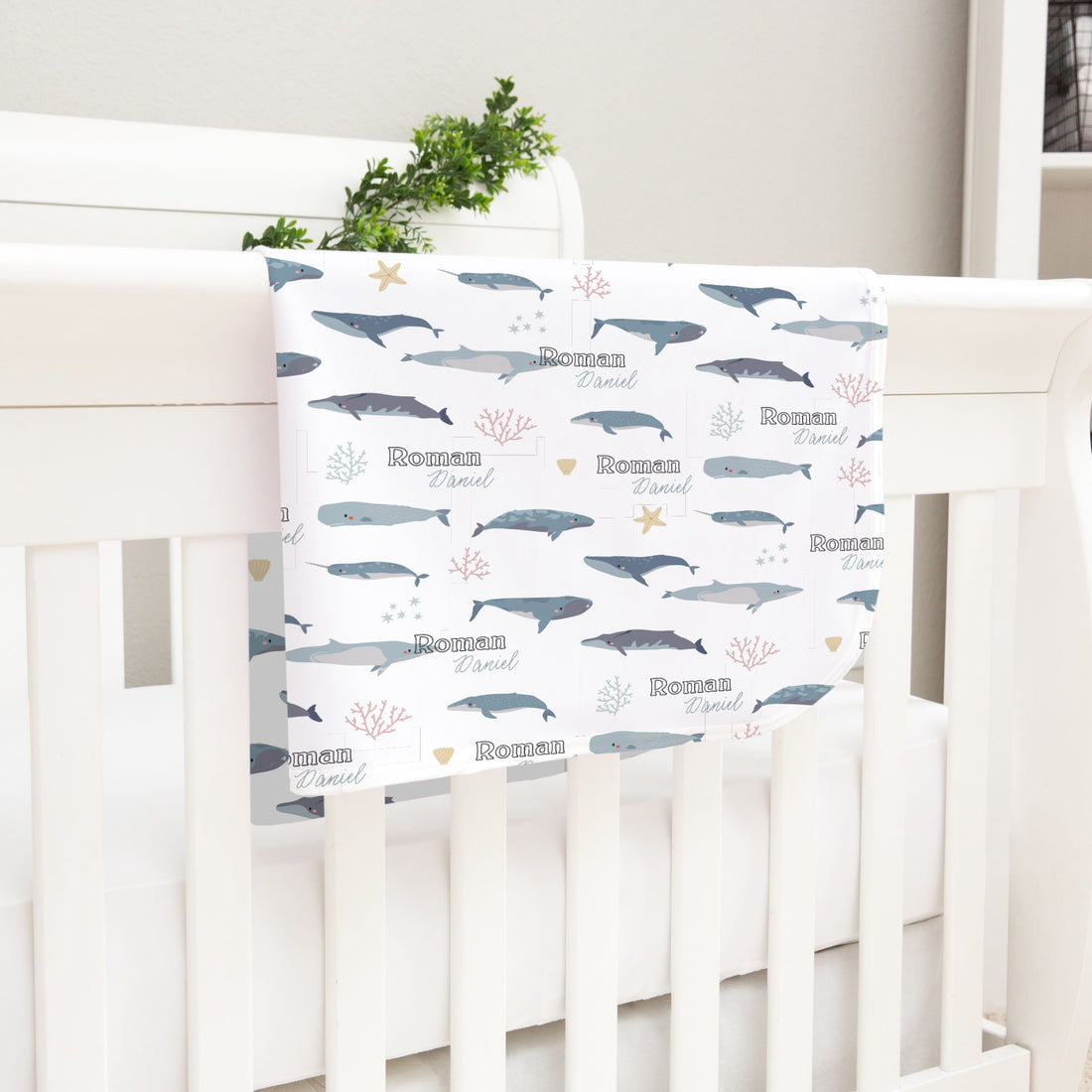 A white crib with a white blanket featuring a repeating pattern of blue and gray whale illustrations, along with the names "Roman" and "Daniel" written on it.
