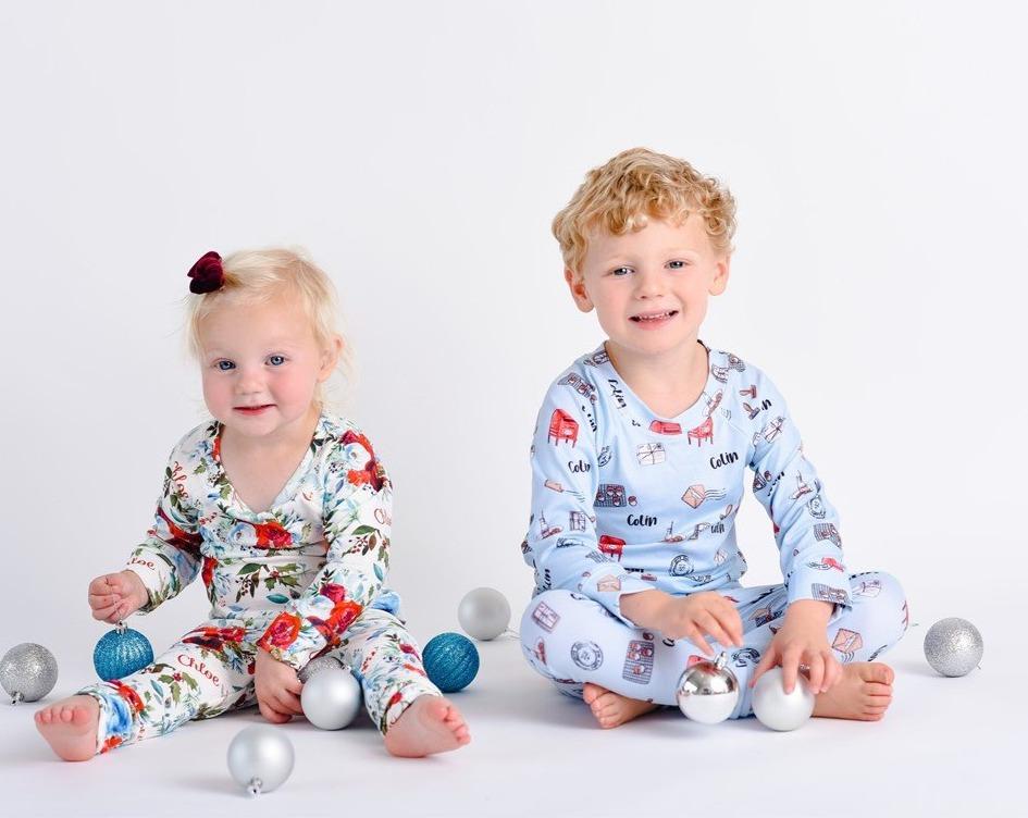 Baby girl wearing Floral Christmas Pajamas with bouquets of red, white, and light blue flowers and holly leaves, and sitting next to her brother in Christmas pajamas. 