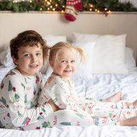 Two siblings sitting on a bed wearing matching Gingerbread Christmas Pajamas. 